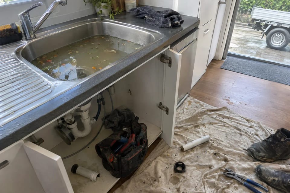 Before: a stainless kitchen sink full of grey standing water with food scraps, under-sink cupboard open.