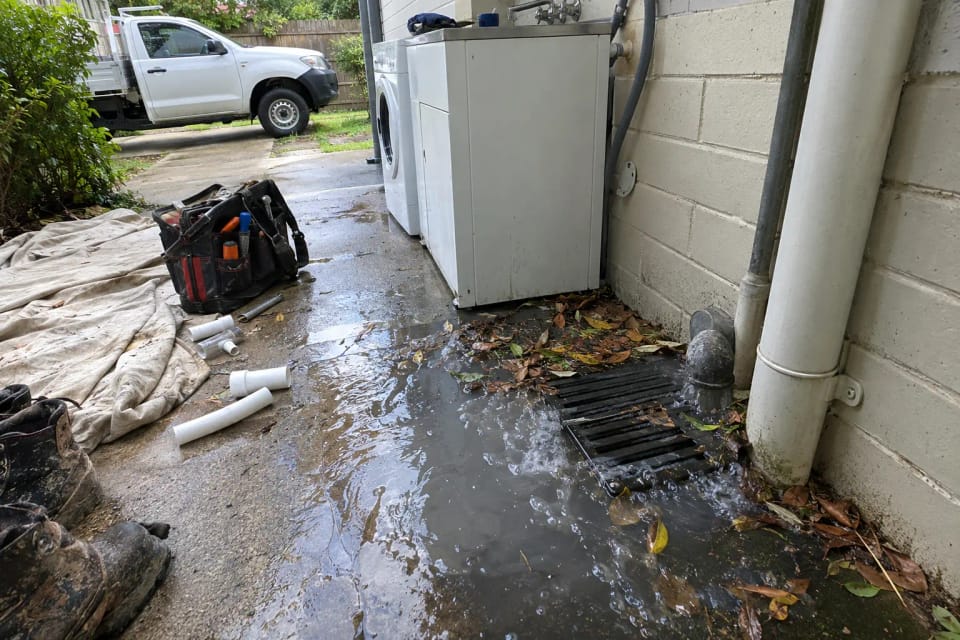 Before: an outdoor overflow gully beside a laundry wall overflowing with grey water, leaves around the grate.